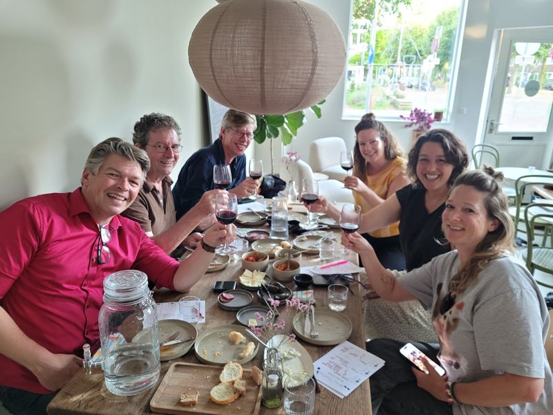 Zeven gasten proosten met rode wijn aan de lange tafel, met mezze en score cards voor zich
